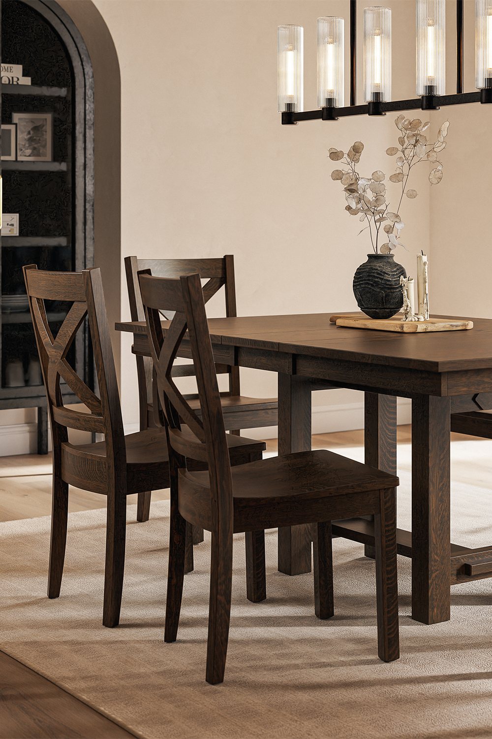 A dark-stained maple wood dining table with matching X-back chairs, set in a warmly lit dining room with a modern chandelier and neutral decor.
